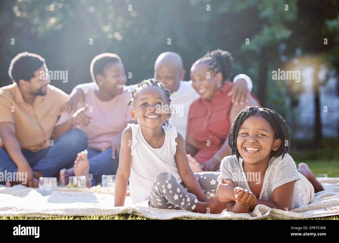 Los niños felices, el picnic y la familia negra se relajan, se unen y disfrutan del tiempo libre juntos en la naturaleza. Retrato de pequeñas hermanas africanas sonríen en felicidad para el verano al aire libre con los padres y los abuelos Foto de stock