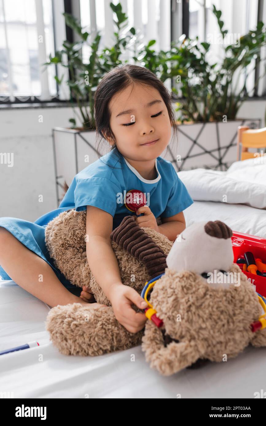 chica asiática sonriente en bata de hospital jugando con oso de peluche y  estetoscopio de juguete en la cama en clínica pediátrica, imagen de stock  Fotografía de stock - Alamy