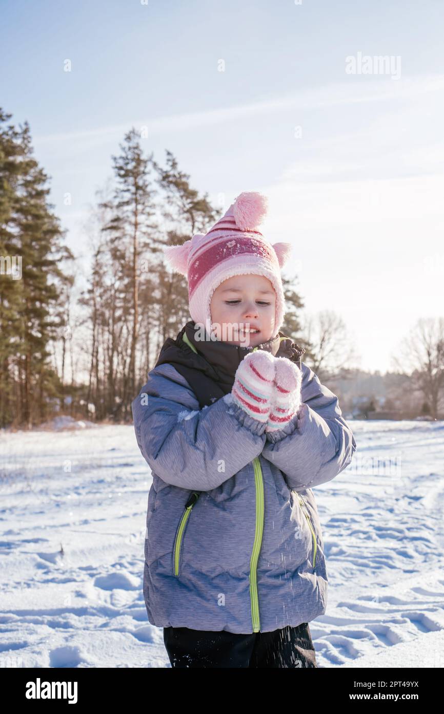 Niña de 3-4 años en el mono de invierno, sombrero y mitones, se para al contra el cielo azul y los abetos arroja nieve sobre su risa Fotografía de