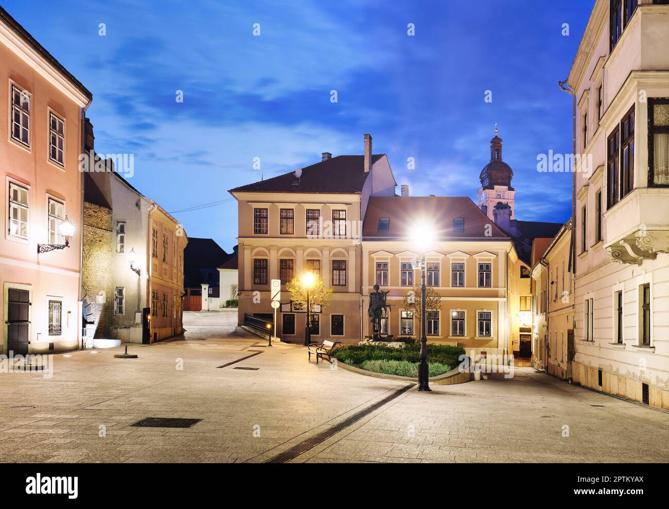 Hungría - Acogedora pequeña plaza barroca en el centro de Gyor, con la escultura de bronce de Nimrod, en el fondo la torre de la catedral por la noche Foto de stock