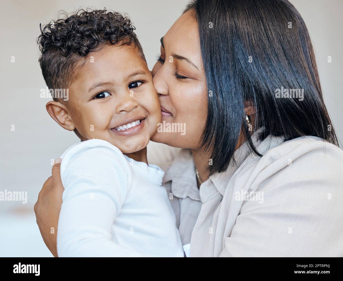Mujer de raza mixta besando a su pequeño hijo adorable en la mejilla mientras se unen en casa. Pequeño niño hispano sonriendo y mirando feliz de ser getti Foto de stock