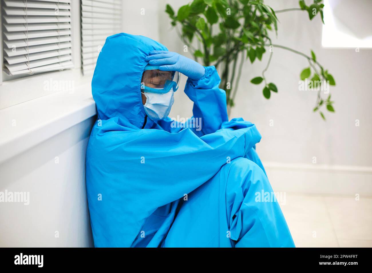 Mujer cansada en equipo de protección personal PPE Sentirse deprimido después de un difícil cambio estresante durante la pandemia de Covid-19, sentada en el suelo Foto de stock