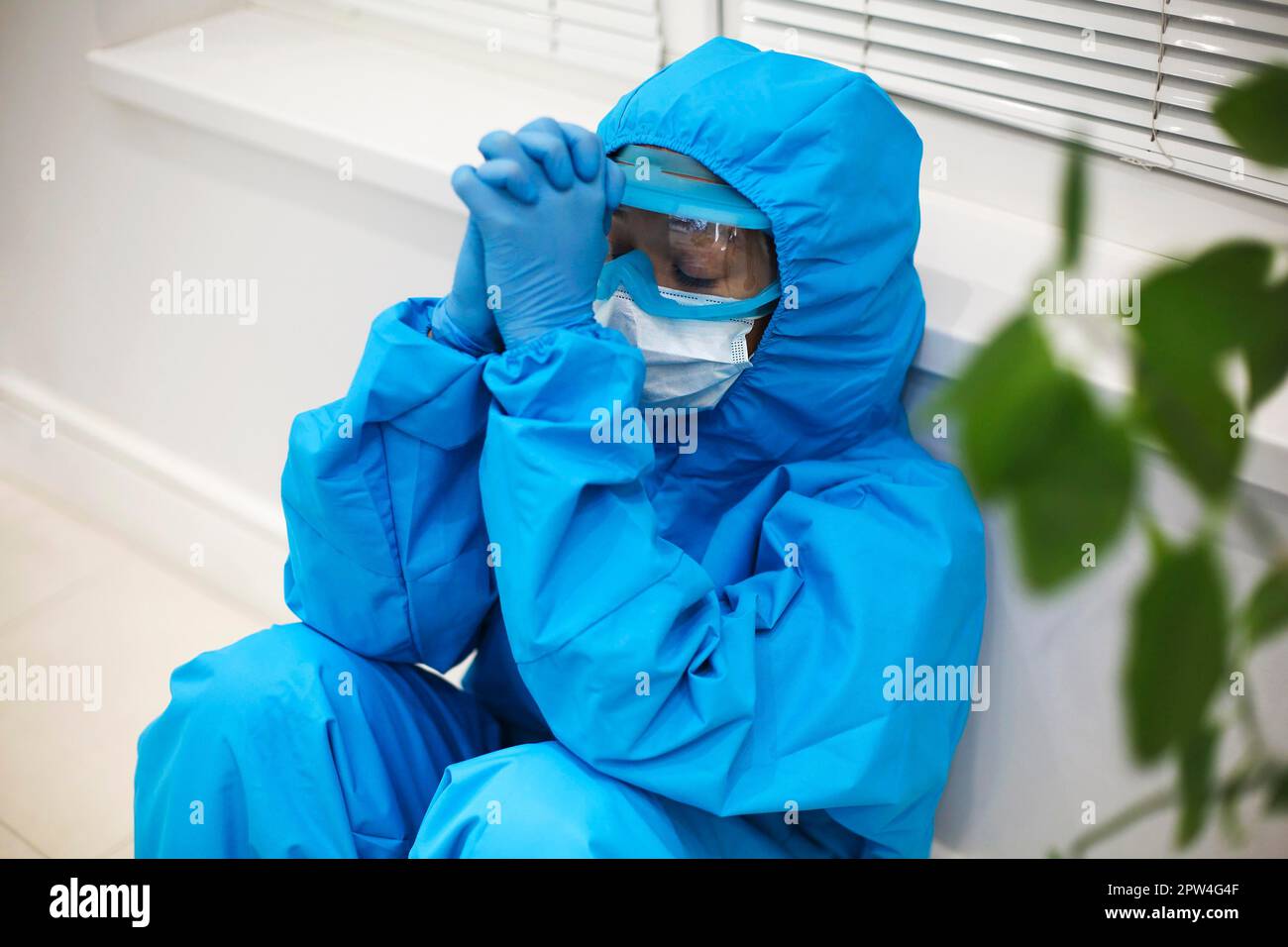 Mujer cansada en equipo de protección personal PPE Sentirse deprimido después de un difícil cambio estresante durante la pandemia de Covid-19, sentada en el suelo Foto de stock