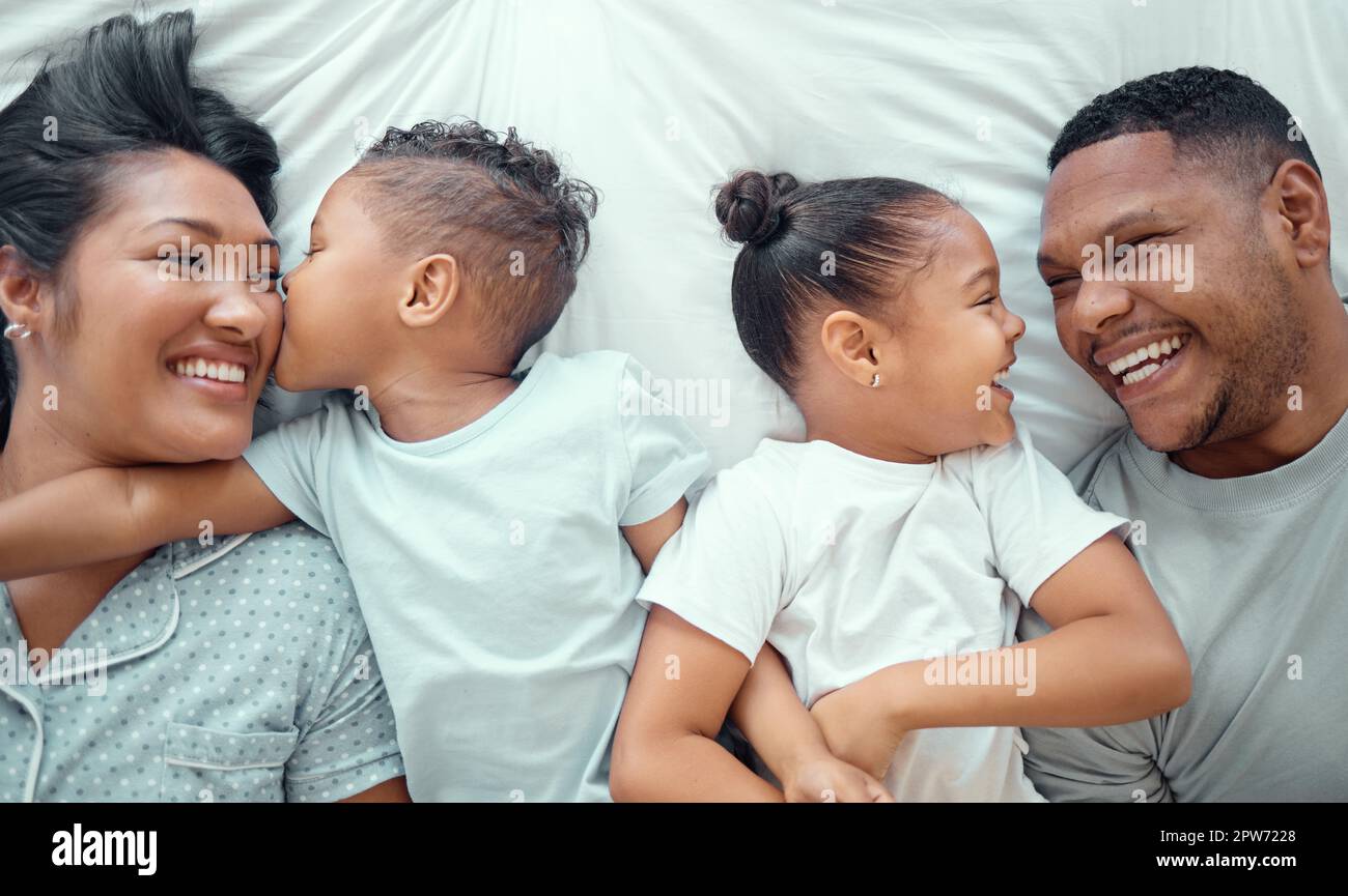 Familia de raza mixta divertida feliz con dos niños relajándose y acostándose juntos en una cama en casa, desde arriba. Adorable niño besando a su madre en th Foto de stock
