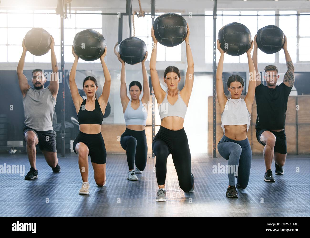 Grupo diverso de jóvenes activos haciendo ejercicios de lunge con balón de medicina aérea mientras entrenan juntos en un gimnasio. Atletas enfocados que desafían los temblos Foto de stock