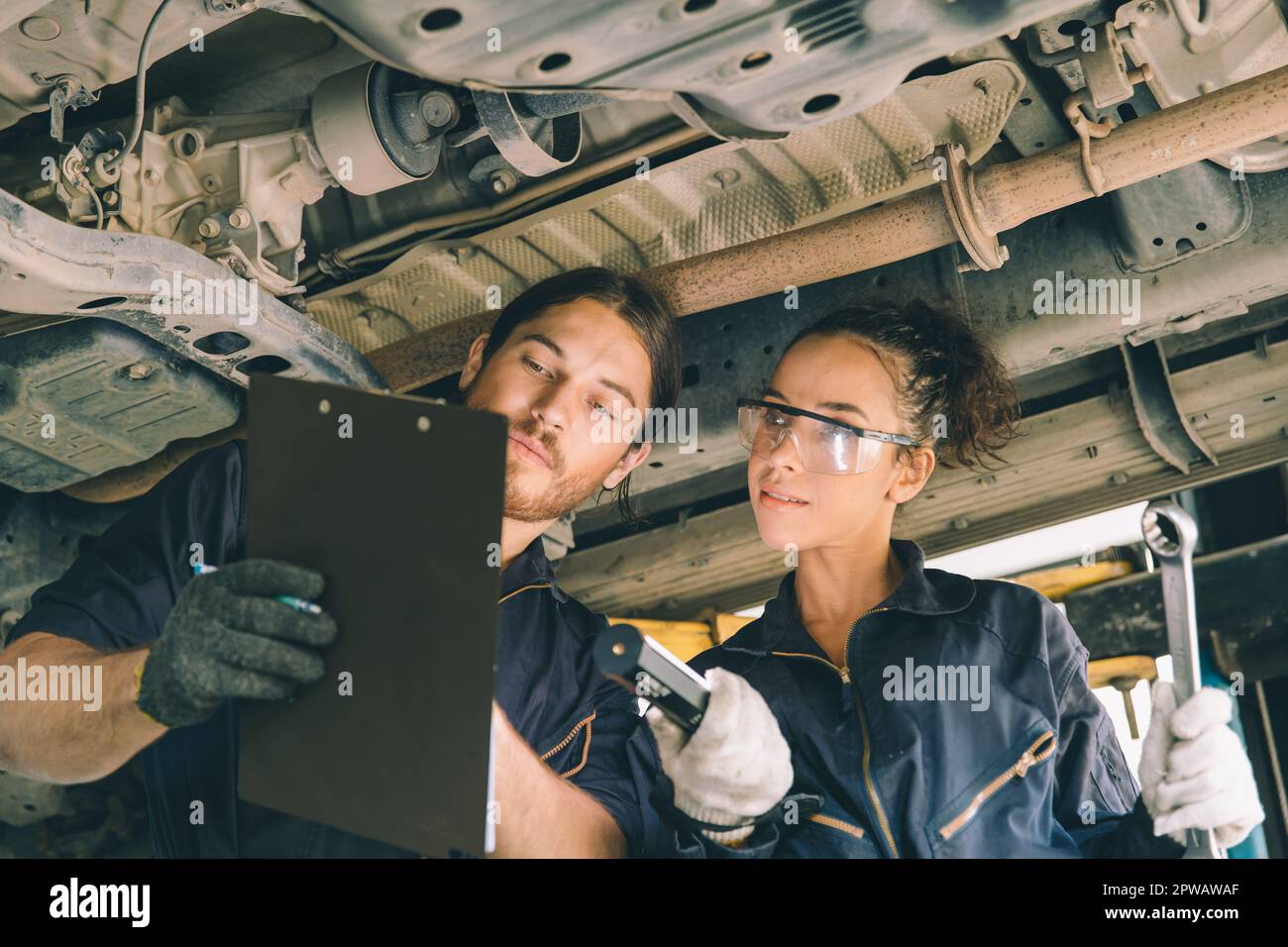 Hombre y mujer del equipo de servicio mecánico que trabajan juntos debajo del alzamiento del coche que comprueba el mantenimiento de la suspensión de la lista en el wokshop auto del garaje Foto de stock