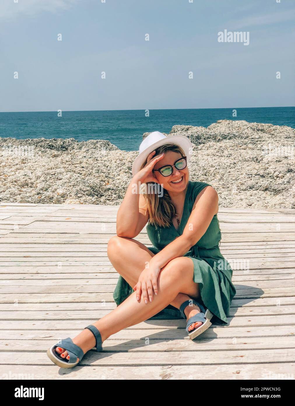 Mujer alegre feliz que lleva gafas de sol y sombrero que disfruta del viaje de vacaciones de verano en la playa, mujer sonriente que se sienta en la terraza de madera en la playa en soleado Foto de stock