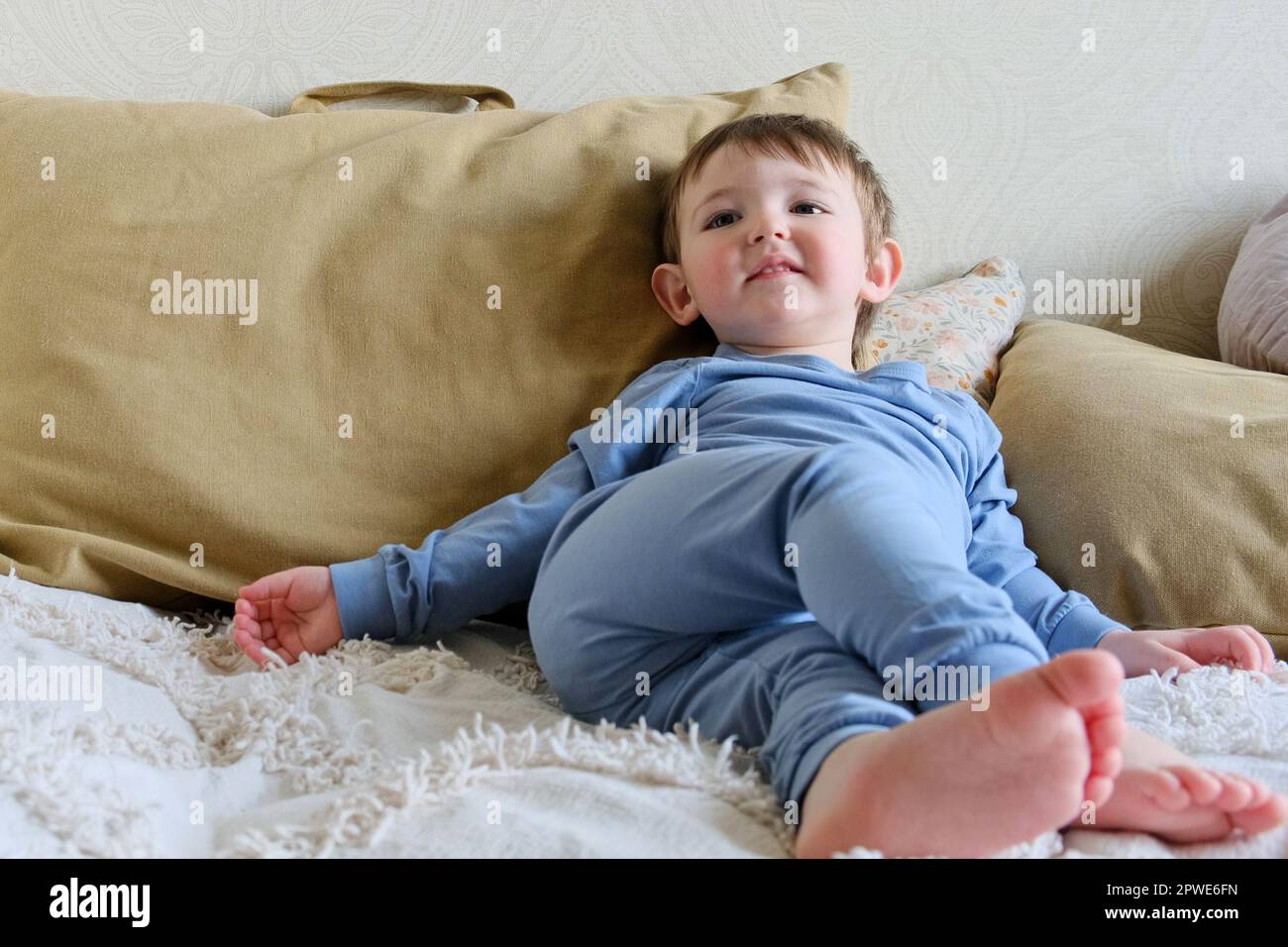Bebé en una ropa azul iaper se sienta en un sofá y mira la televisión. El  niño descansa sentado en un pijama en una cama con almohadas. Niño de unos dos  años
