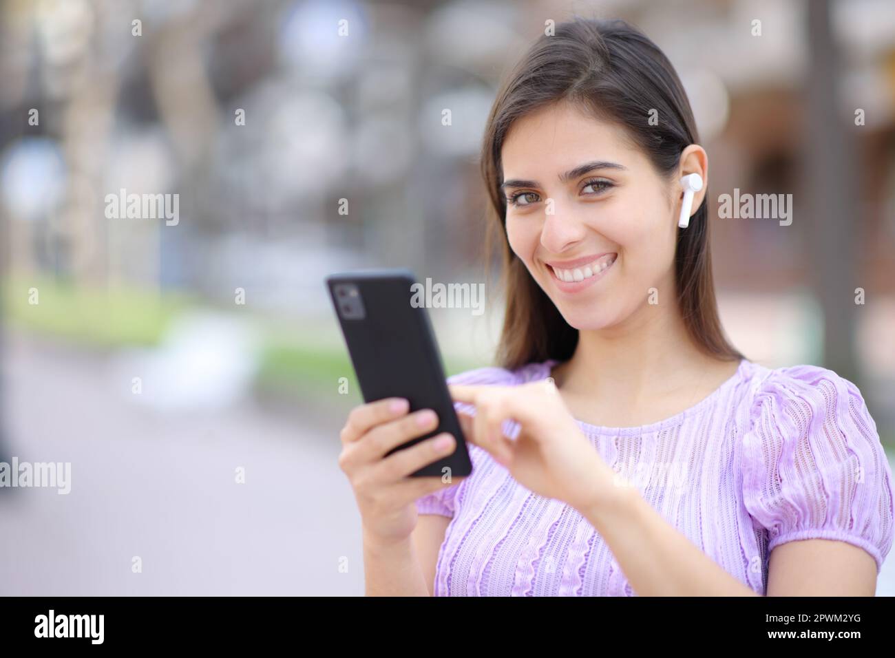 Mujer feliz posando mirando a la cámara con el auricular y el teléfono en la calle Foto de stock