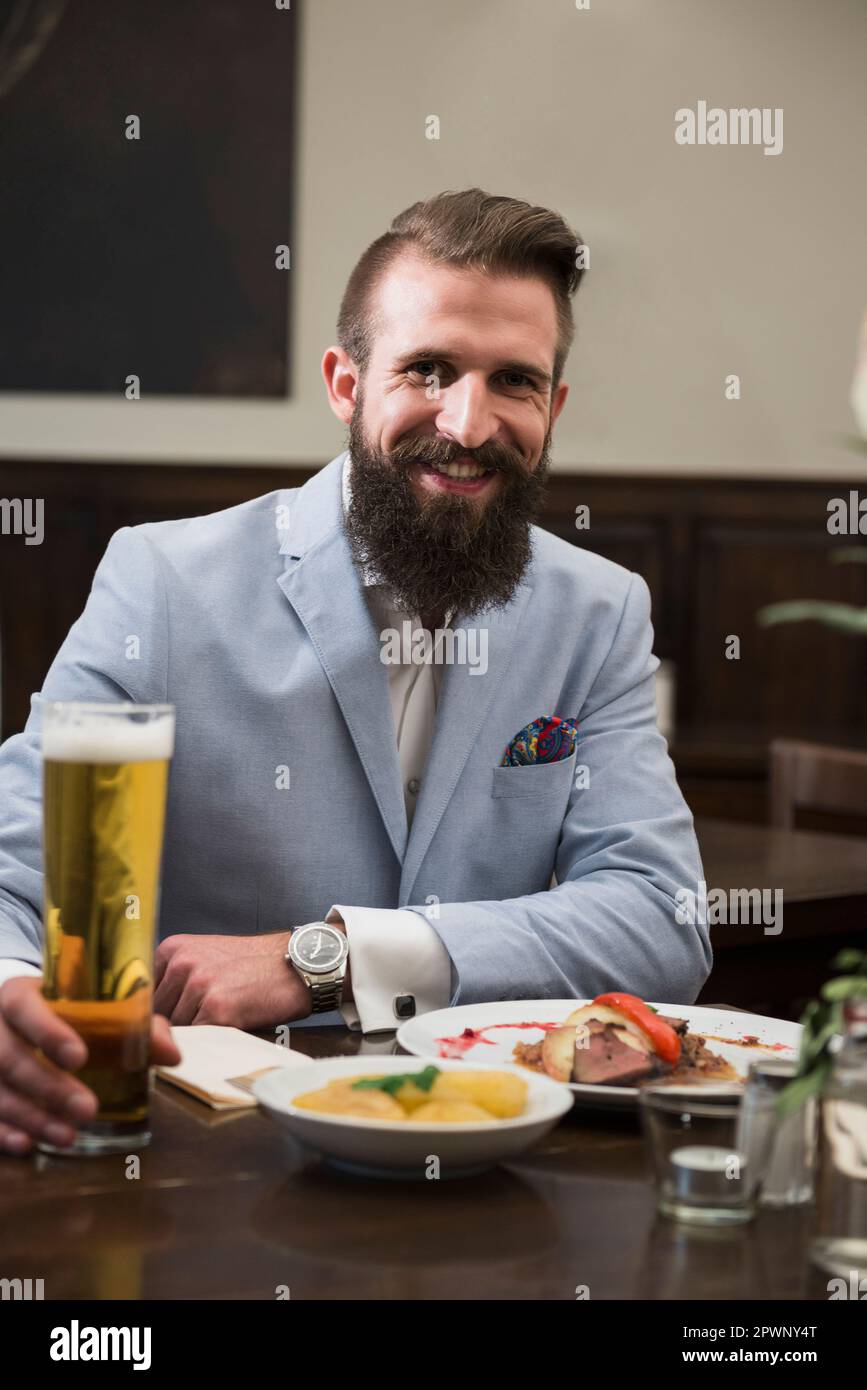 Hombre bien vestido sosteniendo vaso de cerveza y comiendo comida en el restaurante Foto de stock