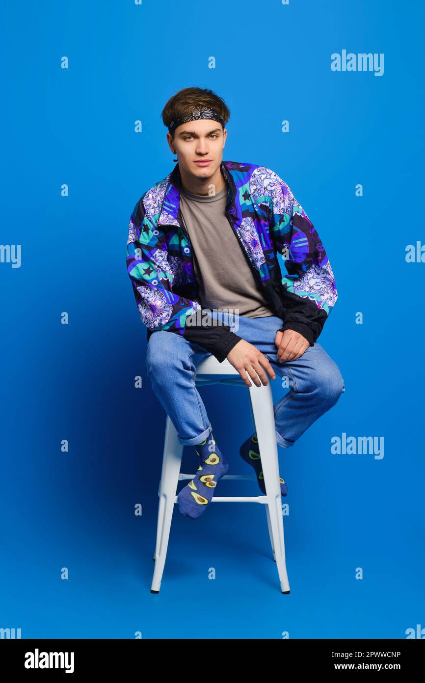 Hombre joven con chaqueta cortaviento, camisa de olivo, jeans calcetines con estampado se sienta en silla alta sobre fondo azul de estudio Fotografía de stock - Alamy