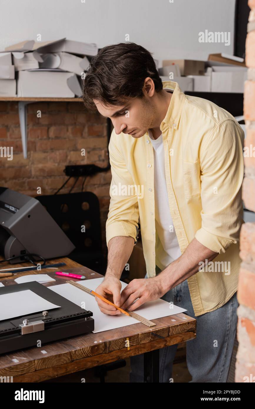 operador de impresión de buen aspecto que sostiene la regla y el lápiz cerca del papel en el centro de impresión moderno, imagen de stock Foto de stock