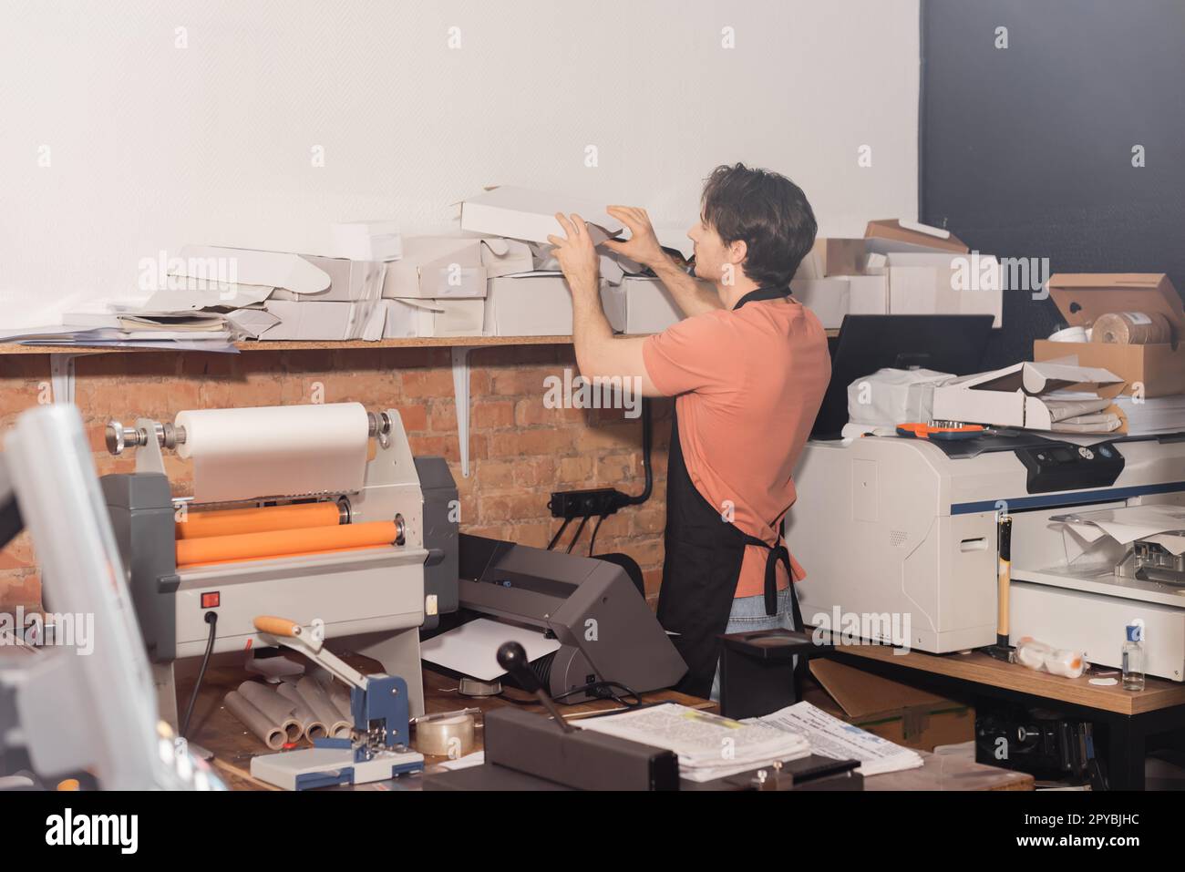 joven tipógrafo en delantal alcanzando cajas de cartón dobladas junto al equipo en el centro de impresión, imagen de stock Foto de stock