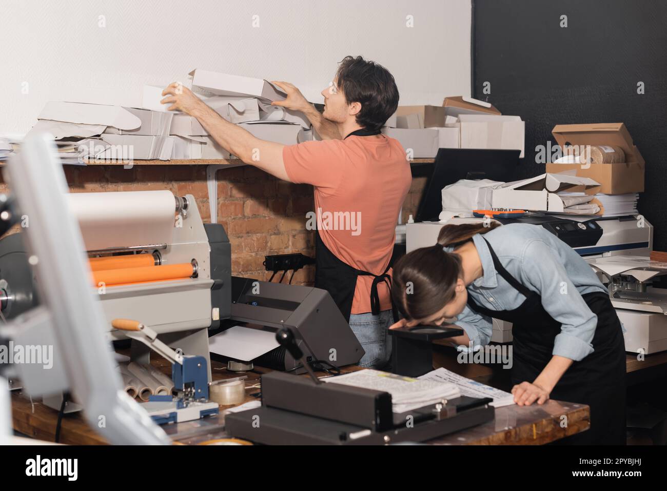 tipógrafo en delantal alcanzando cajas de cartón plegadas junto a colega y equipo en el centro de impresión, imagen de stock Foto de stock
