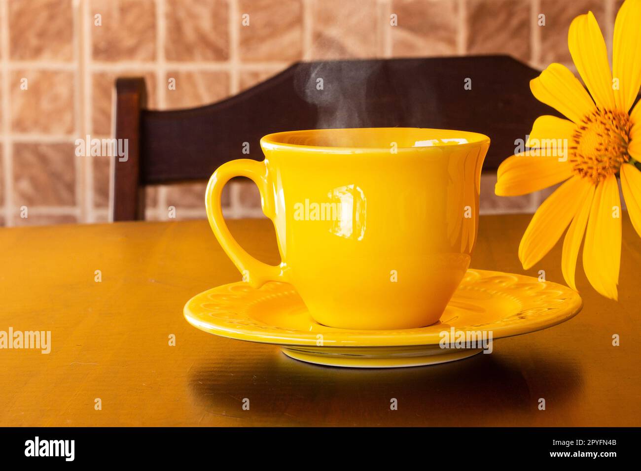 Goiânia, Goias, Brasil – 01 de mayo de 2023: Una taza amarilla llena de café, con un platillo y un girasol, sobre una mesa de madera con respaldo de silla. Foto de stock