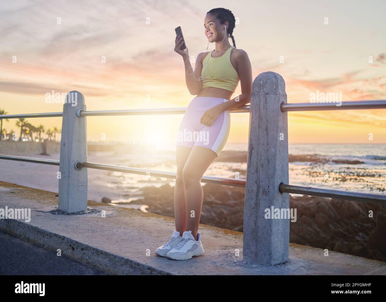 Fitness, playa y mujer con smartphone después de correr al atardecer, música en línea y streaming o podcast en la naturaleza para hacer ejercicio. Corra, relájese y desplácese por las redes sociales después de correr, sonría con los auriculares. Foto de stock