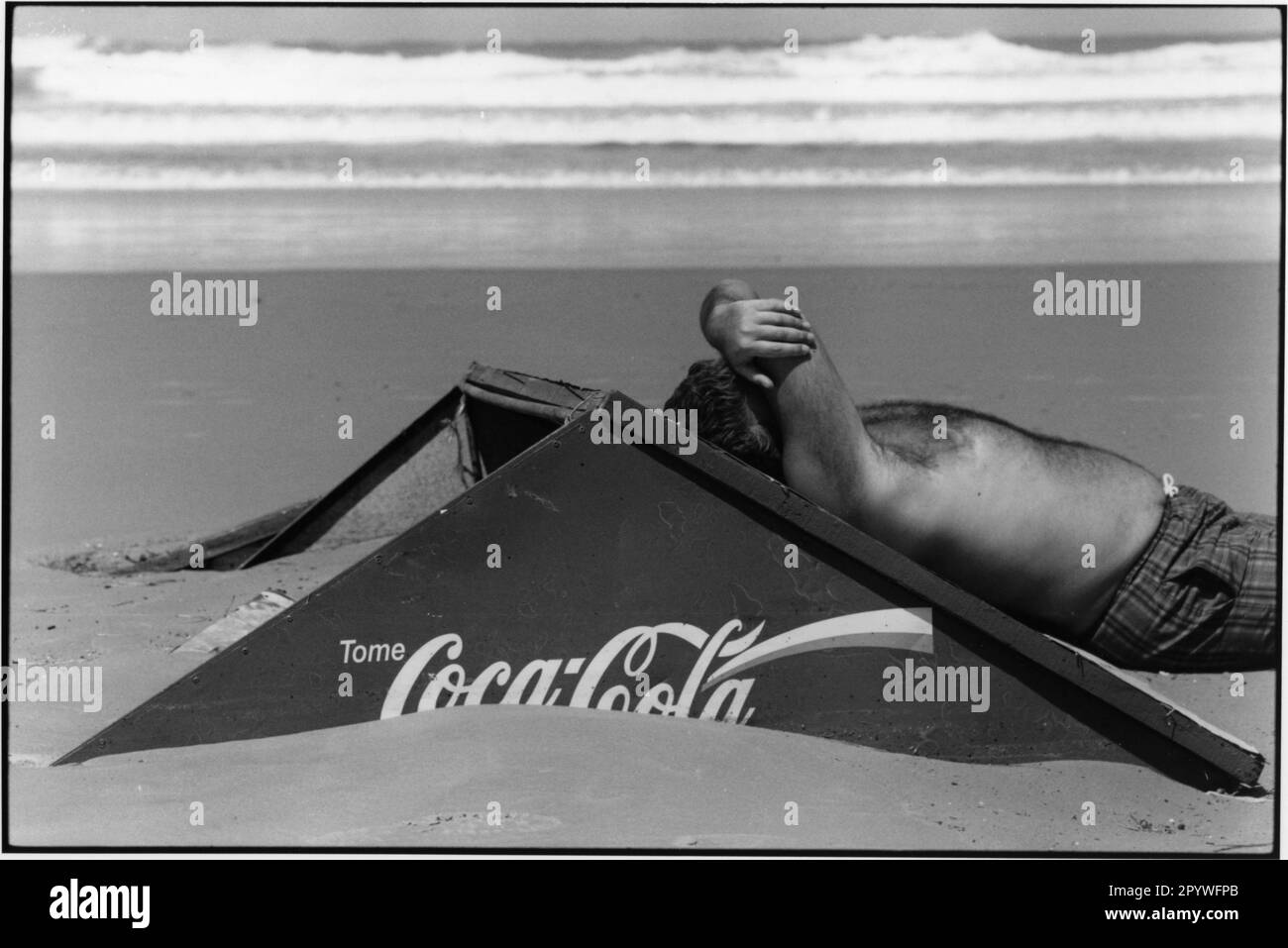 Uruguay, Punta del Este. Tomar el sol. Viaje y tiempo libre: Un hombre se inclina contra una cartelera de Coca-Cola en la playa, con las olas del Atlántico en el fondo. Escena de playa, blanco y negro. Foto, 1997. Foto de stock
