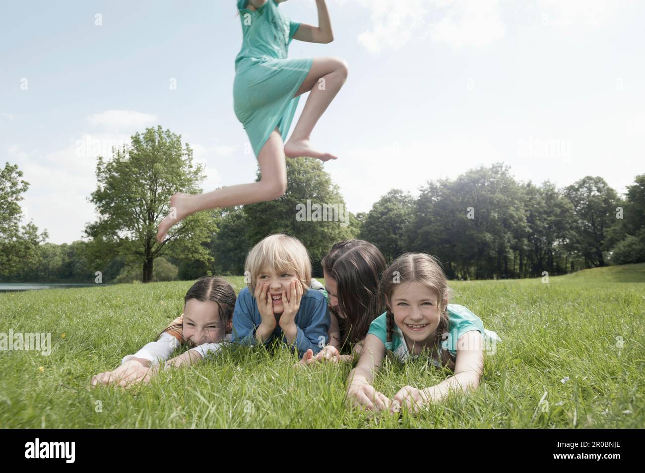 Chica saltando sobre sus amigos tumbados en la hierba en un parque, Munich, Baviera, Alemania Foto de stock
