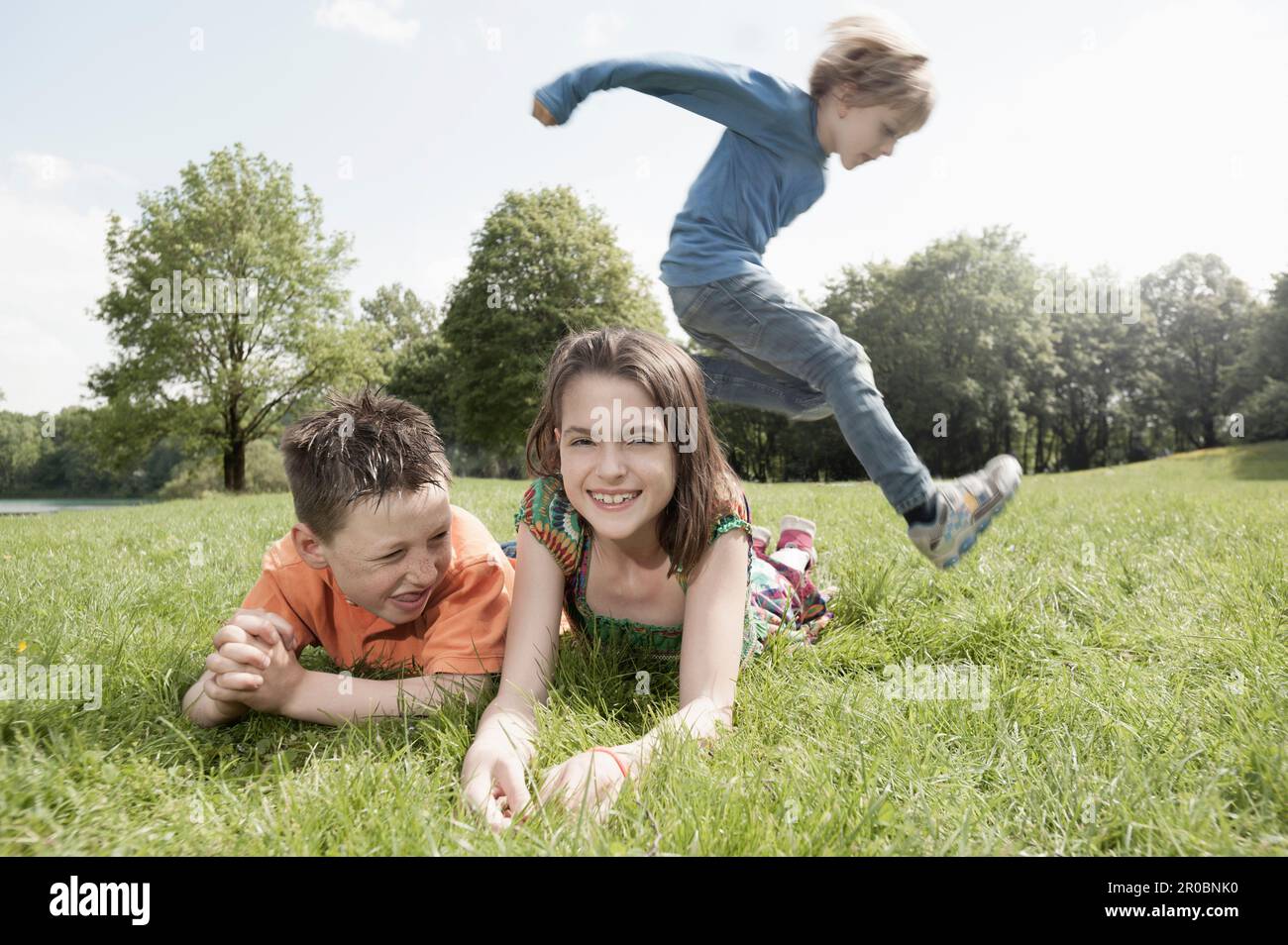 Niño saltando sobre sus amigos tumbados en la hierba en un parque, Munich, Baviera, Alemania Foto de stock