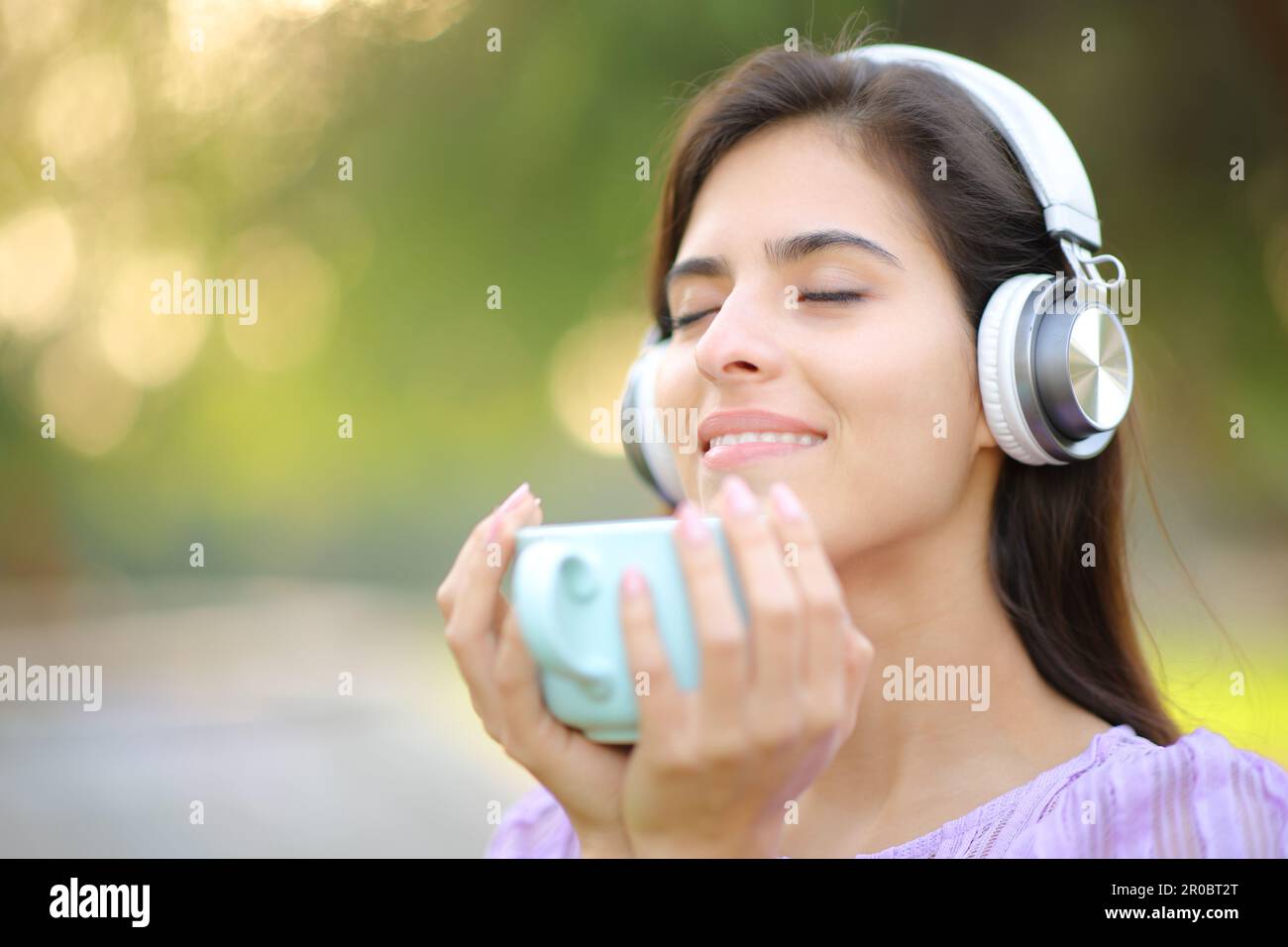 Mujer relajada que lleva auriculares escuchando música y bebiendo café en un parque Foto de stock