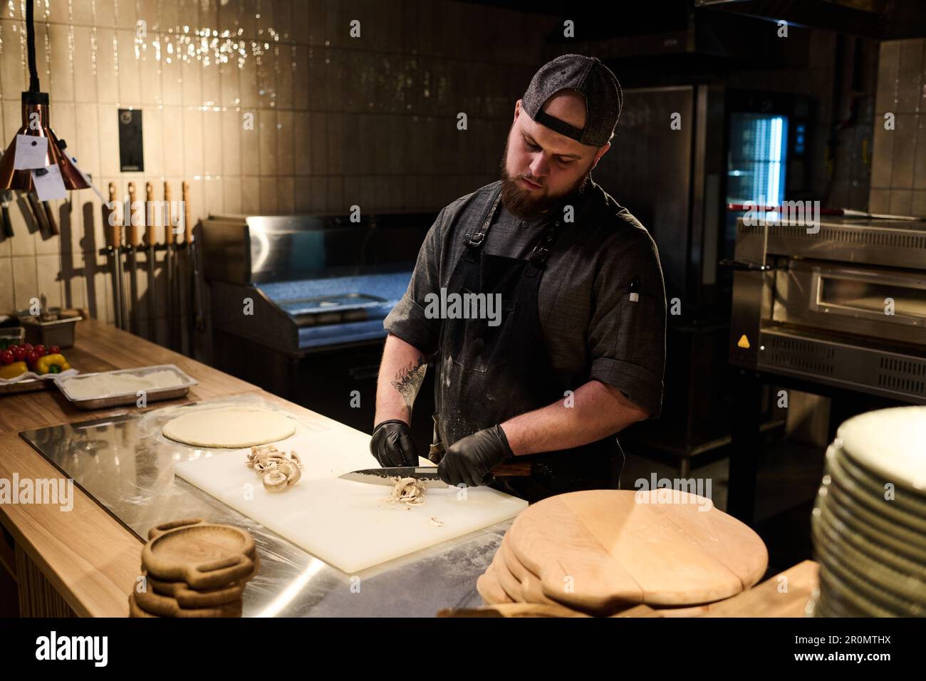 Buzo Alinear animal Joven chef con guantes negros y delantal picando champiñones frescos en  tabla de cortar mientras prepara pizza por mesa en la cocina Fotografía de  stock - Alamy