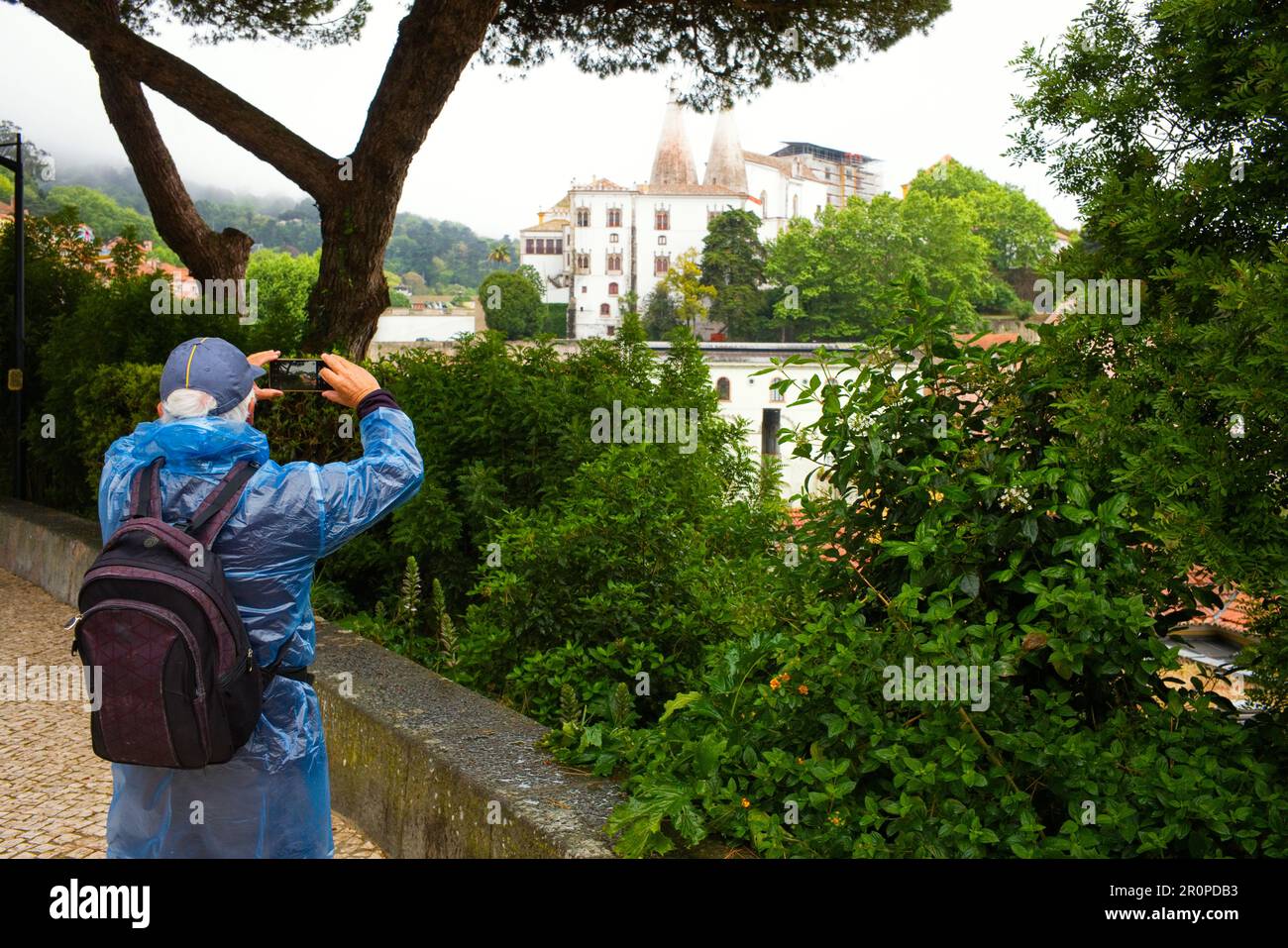 Un turista que lleva una cubierta de plástico para la lluvia toma una foto del Palacio Nacional de Sintra en un día lluvioso Foto de stock