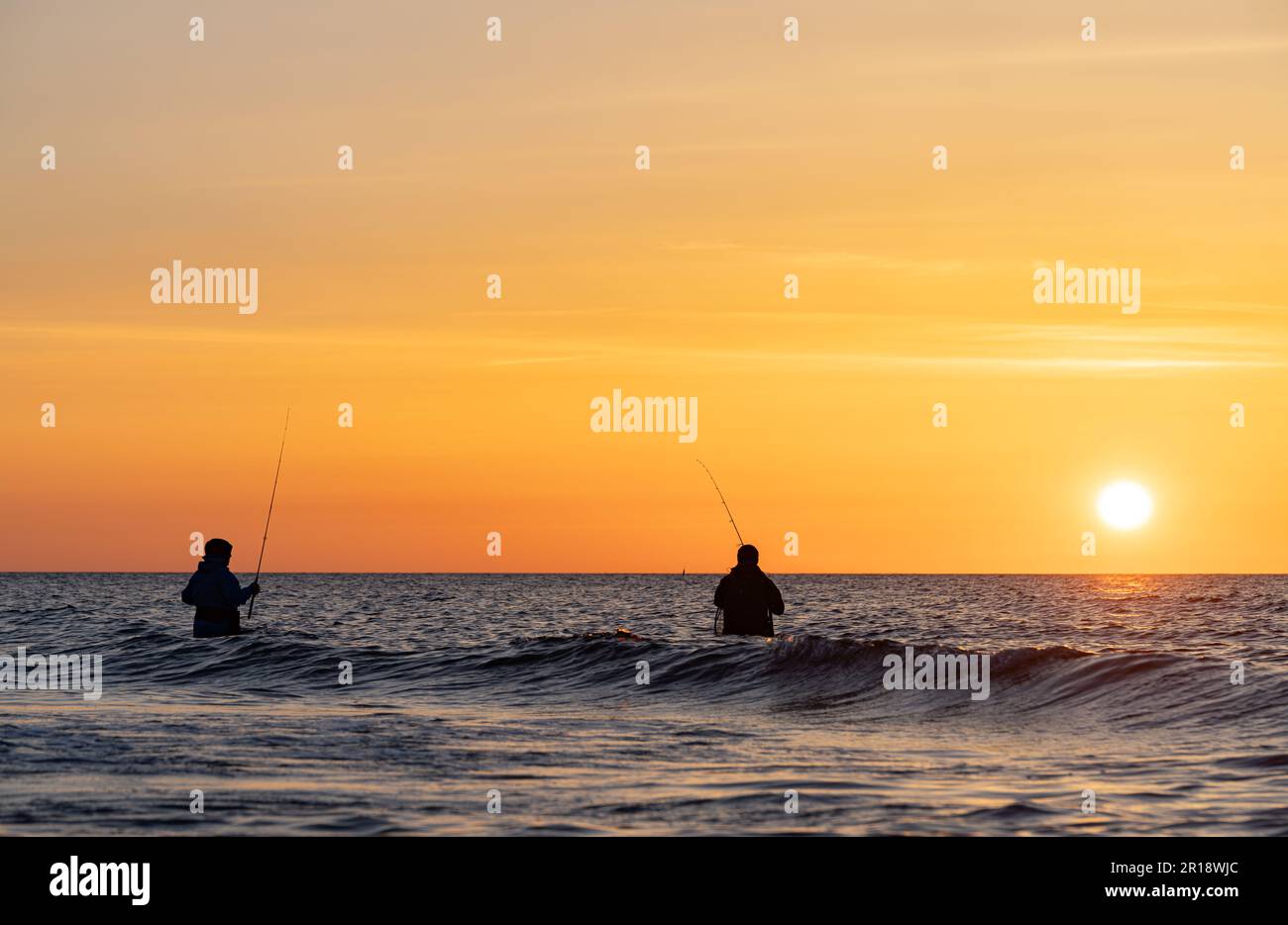 Las siluetas de dos pescadores en pantalones impermeables pescando frente a una puesta de sol naranja de ensueño en el mar Báltico. Están de pie en el medio de Foto de stock