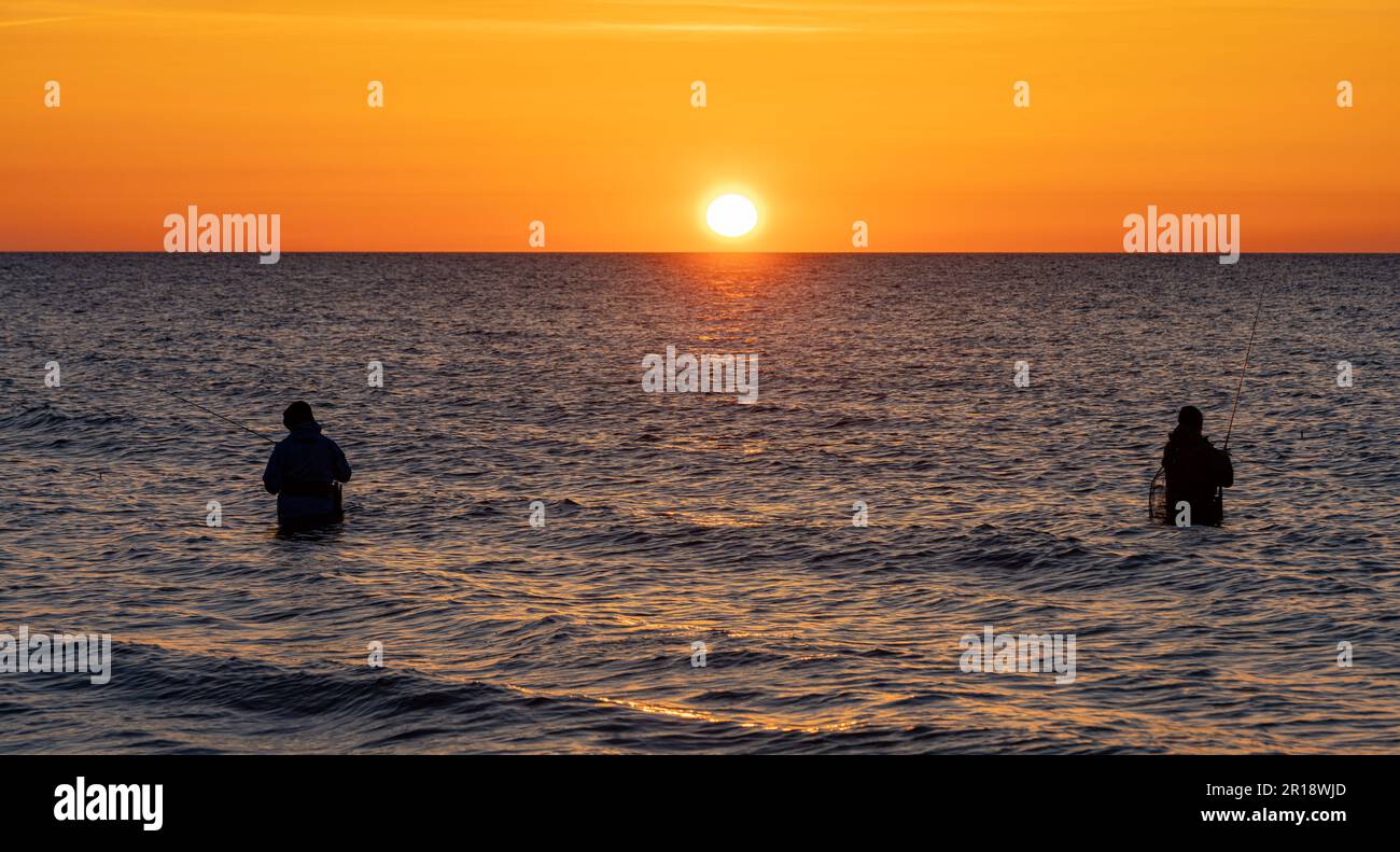 Las siluetas de dos pescadores en pantalones impermeables pescando frente a una puesta de sol naranja de ensueño en el mar Báltico. Están de pie en el medio de Foto de stock
