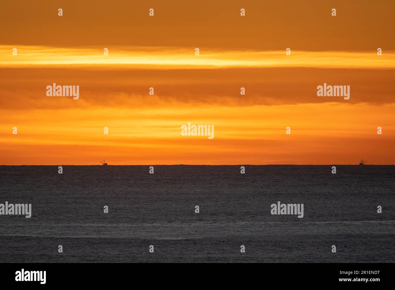 Atardecer cielo naranja sobre el Océano Pacífico, con dos pequeños barcos de pesca en el horizonte Foto de stock