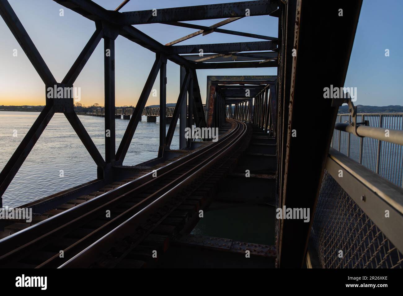 En la silueta retroiluminada en el patrón estructural de la salida del sol del puente del braguero y las líneas principales en el puente del ferrocarril de Tauranga a través del puerto. Foto de stock