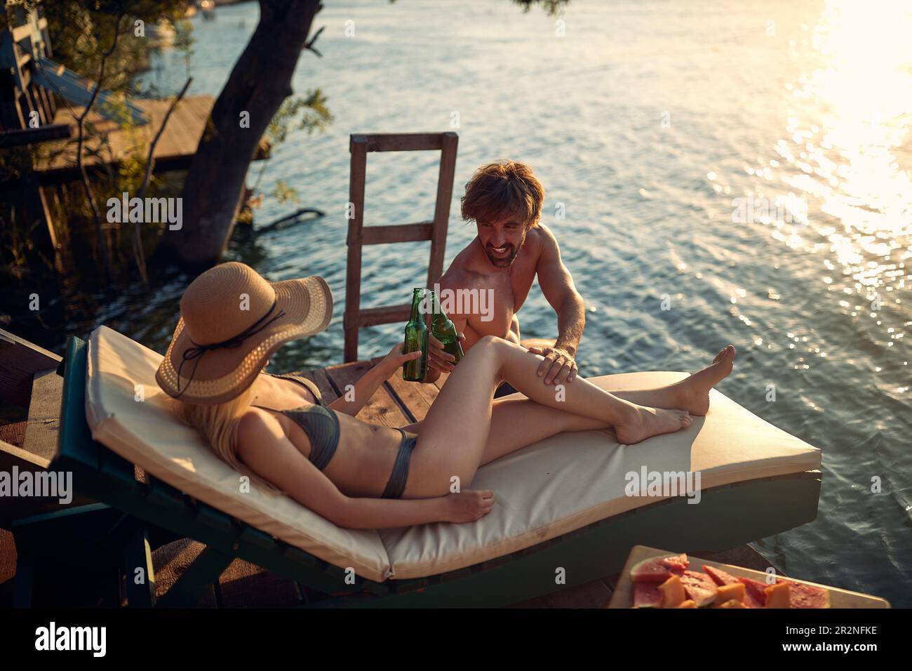 Una joven pareja en traje de baño disfrutando del sol y la bebida en la orilla del río en un hermoso día de verano. Verano, río, vacaciones Foto de stock