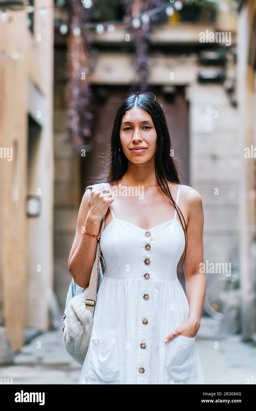 Mujer hispana con mochila sosteniendo la mano en el bolsillo del vestido blanco y mirando a la cámara mientras que está de pie en el fondo borroso de la calle de Barcelona, España Foto de stock