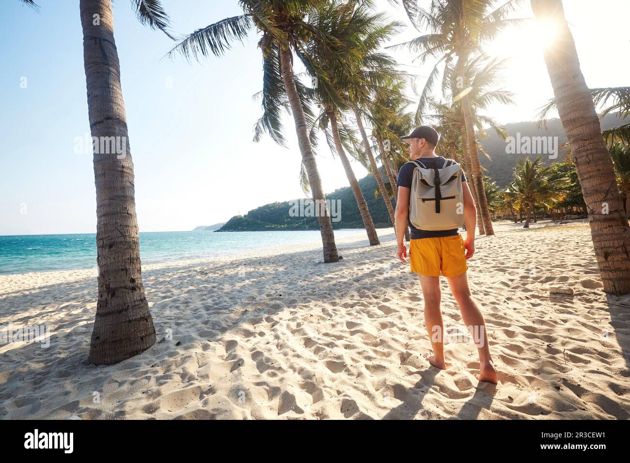 Vista trasera del turista con mochila caminando en la hermosa playa de arena con palmeras y mirando la puesta de sol. Islas Cham, Vietnam. Foto de stock