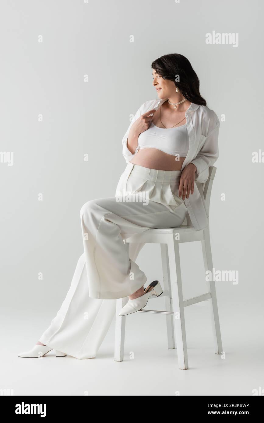 longitud completa de la mujer embarazada de moda en el top blanco de la  cosecha, camisa y pantalones sentados en la silla, sonriendo y mirando  lejos en el fondo gris, fashi de