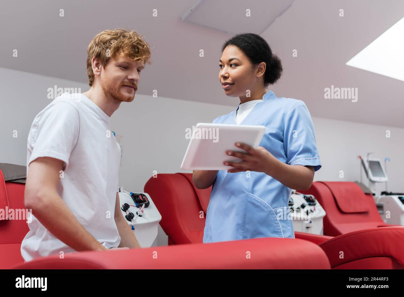 enfermera multirracial en uniforme azul que muestra la tableta digital al donante de sangre pelirroja feliz sentado en una silla médica cómoda cerca de la transfusión automatizada Foto de stock