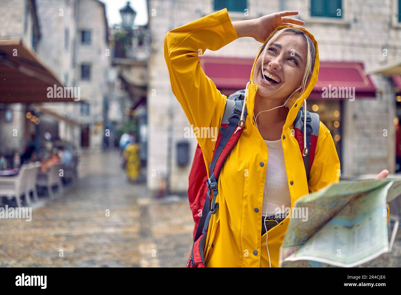 Camina en la ciudad bajo la lluvia. Mujer turística joven en chubasquero con mapa, sintiéndose feliz, sonriendo. Viaje en ciudad nueva. Turismo, estilo de vida, diversión, concepto. Foto de stock