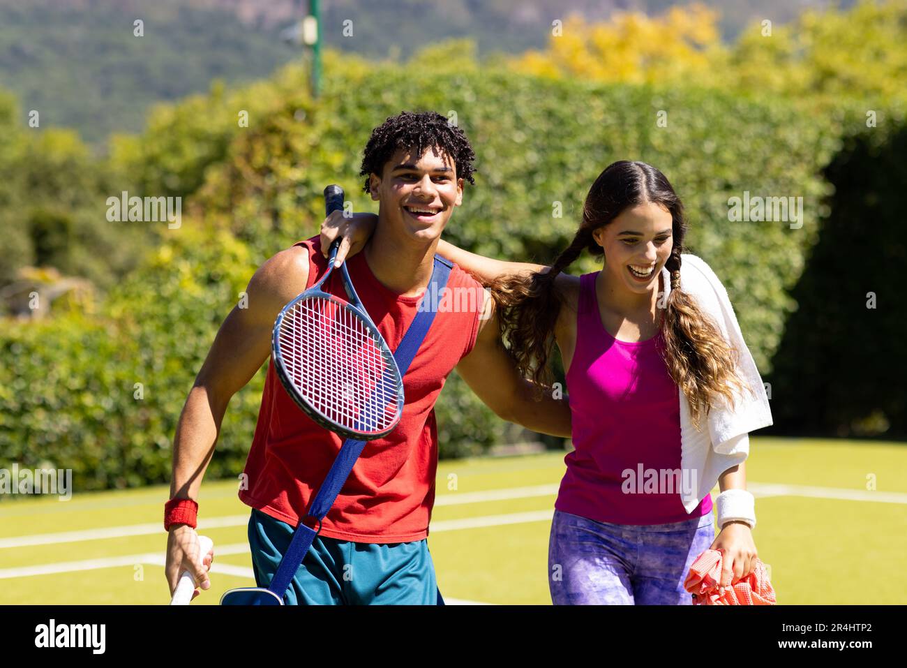 Alegre pareja joven biracial con los brazos alrededor caminando fuera de la cancha de tenis después del partido Foto de stock