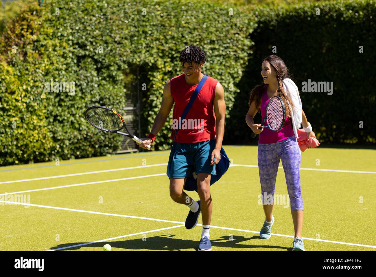 Alegre pareja joven biracial con raquetas saliendo de la cancha de tenis después de jugar Foto de stock
