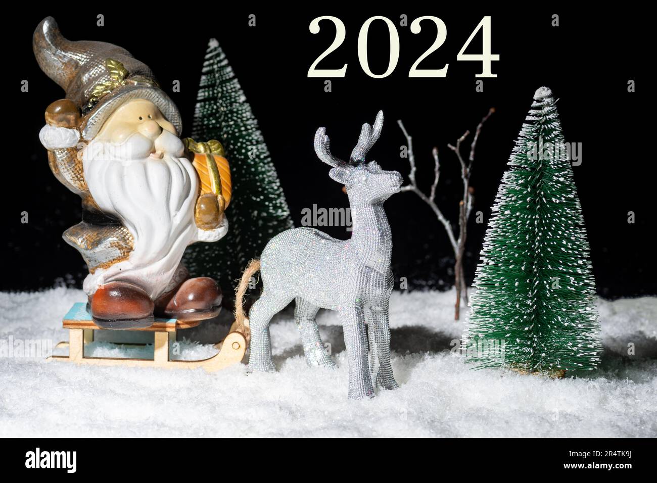 Víspera de Año Nuevo 2024. Un juguete brillante ciervo de plata que lleva un trineo con Santa Claus con regalos a través del bosque nevado entre los árboles de Navidad en la noche Foto de stock