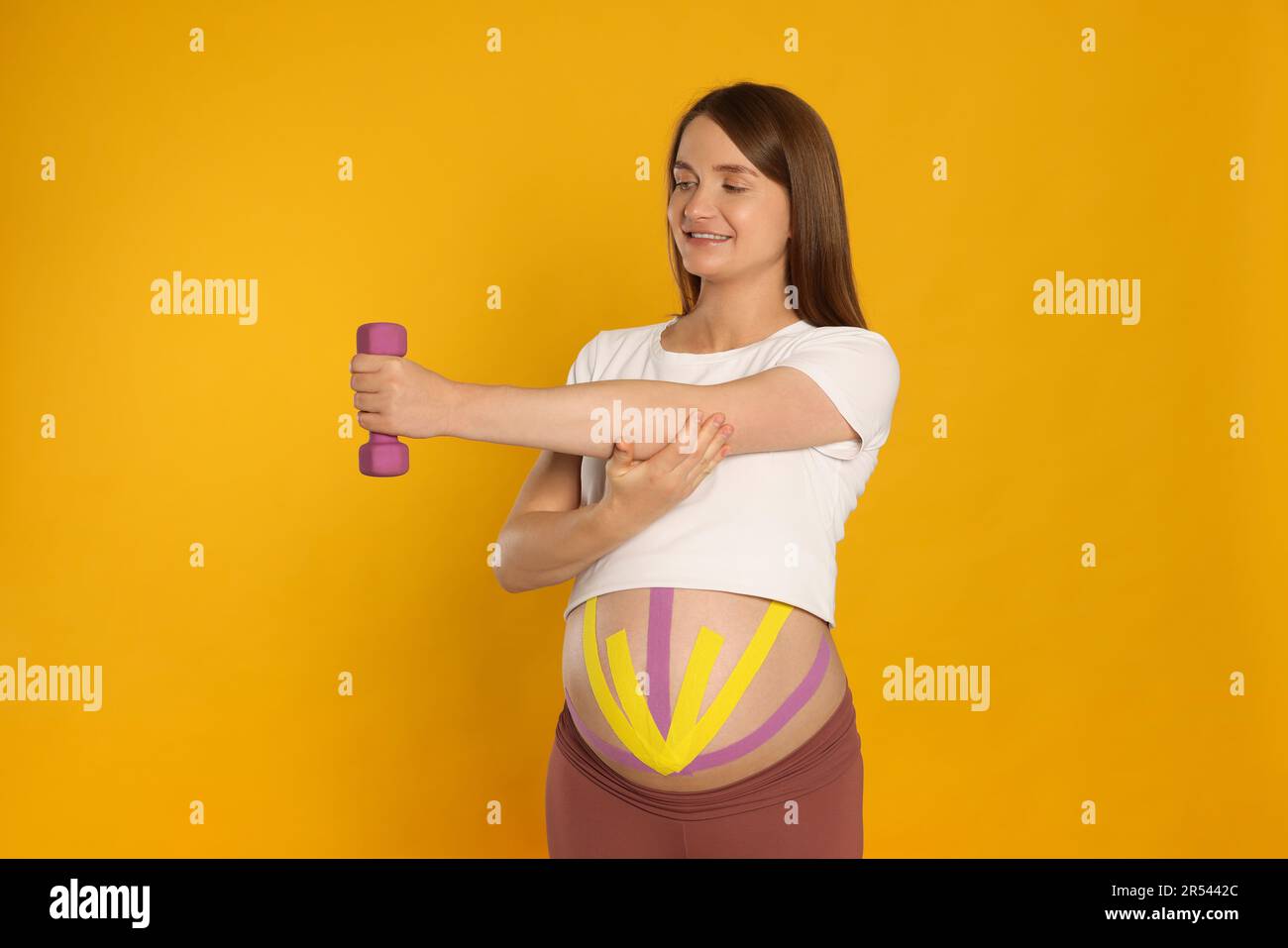 Mujer embarazada con cintas de kinesio en su vientre haciendo ejercicios contra fondo naranja Foto de stock