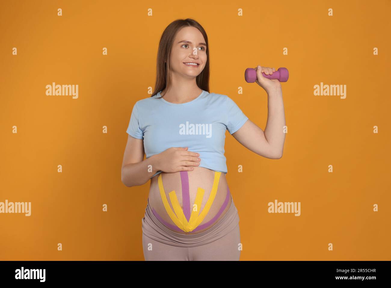 Mujer embarazada con cintas de kinesio en su vientre haciendo ejercicios contra fondo naranja Foto de stock
