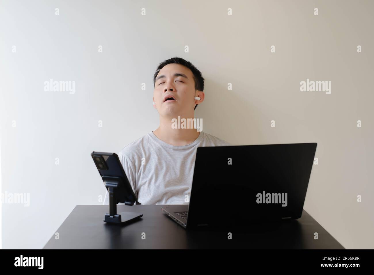 Un joven asiático que lleva una camiseta gris y un par de auriculares inalámbricos se queda dormido mientras trabaja desde casa Foto de stock