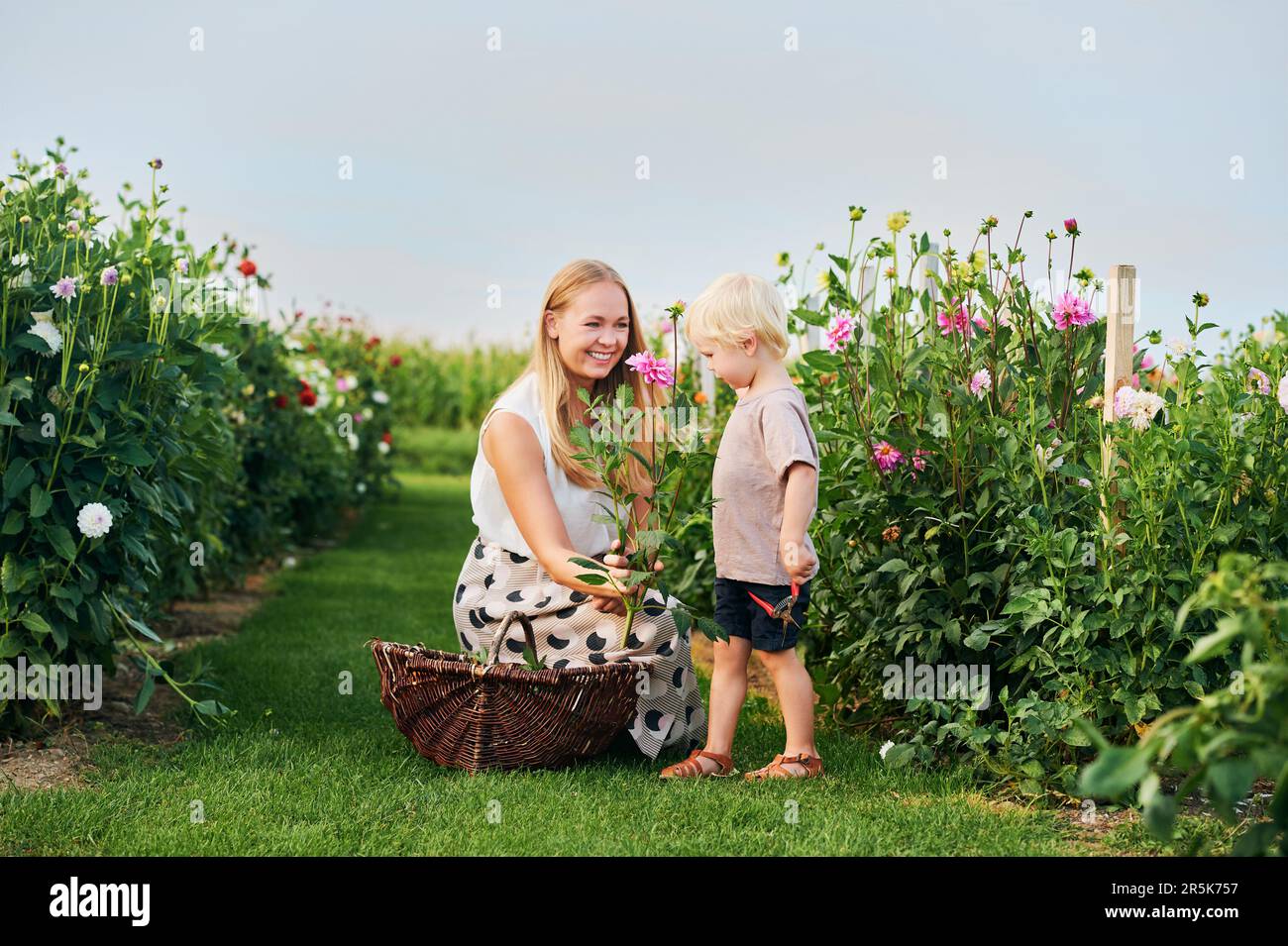 Madre joven feliz y niño pequeño trabajando en hermoso jardín de flores, recogiendo flores, estilo de vida familiar Foto de stock