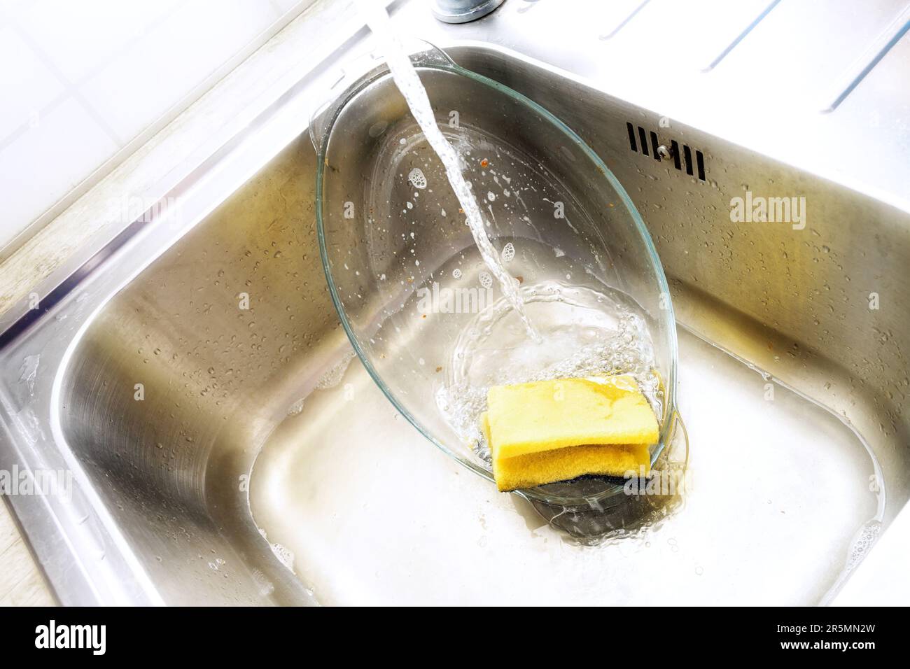 Cazuela de vidrio en el fregadero se limpia con una esponja amarilla y detergente para lavavajillas bajo un chorro de agua, tareas domésticas molestas, espacio de copia, s Foto de stock