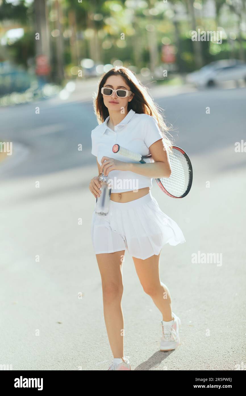 jugador de tenis femenino, mujer joven con el pelo moreno largo que camina  en traje deportivo blanco y zapatos mientras sostiene la raqueta y la  botella con el agua borrosa Fotografía de