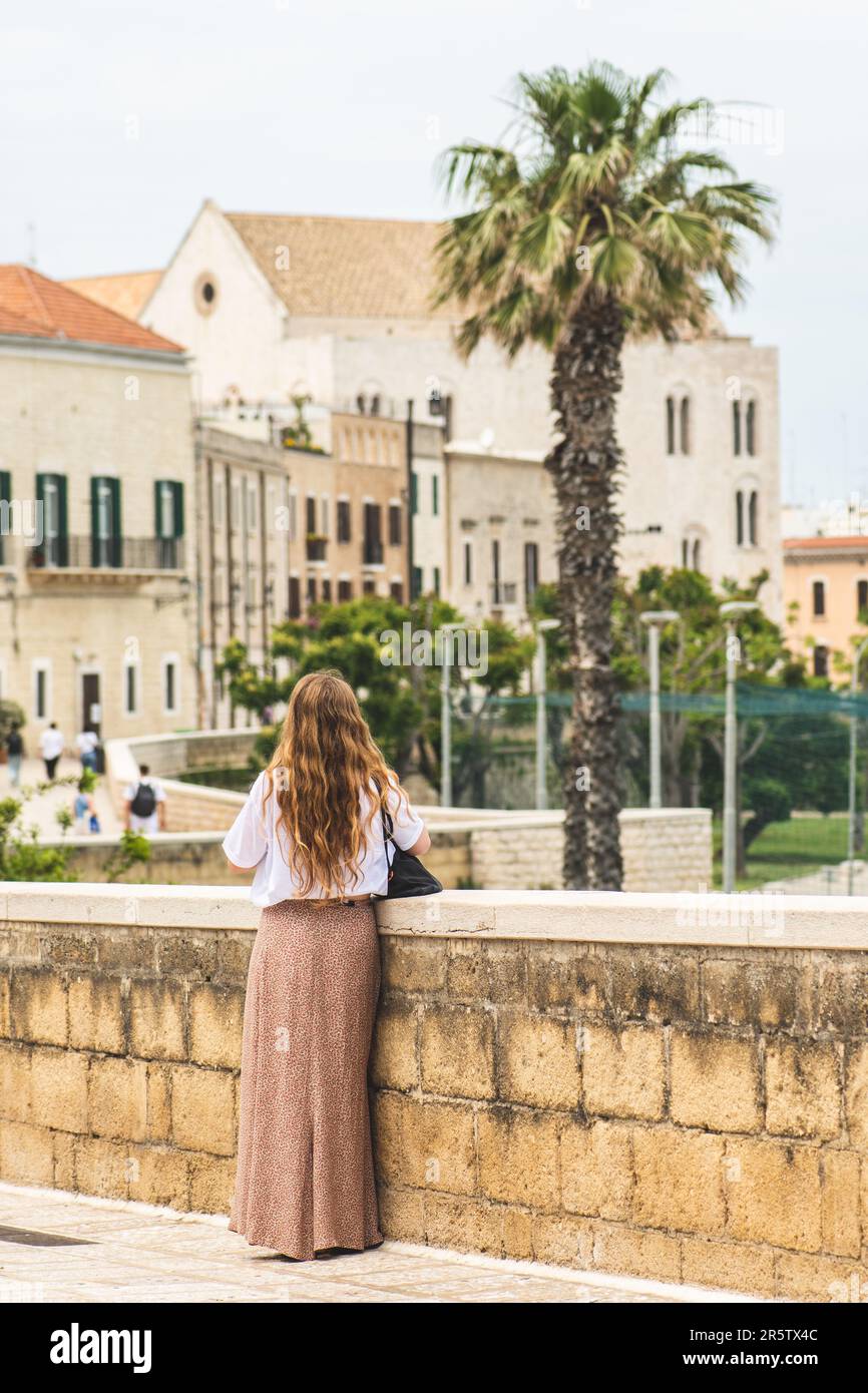 Chica de pelo rojo caminando en las paredes antiguas con la Basílica Pontificia o la Iglesia de San Nicolás y palmeras en el fondo en la ciudad vieja de Bari, Puglia Foto de stock