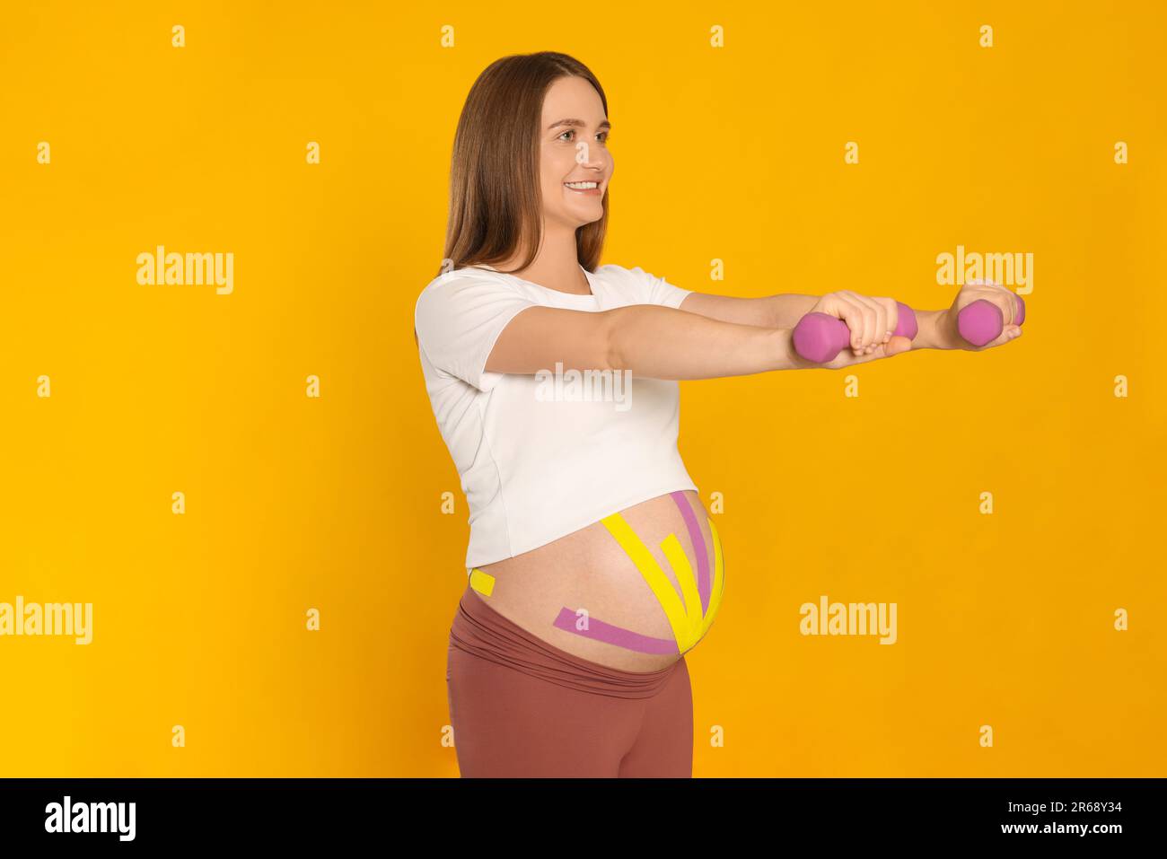 Mujer embarazada con cintas de kinesio en su vientre haciendo ejercicios contra fondo naranja Foto de stock