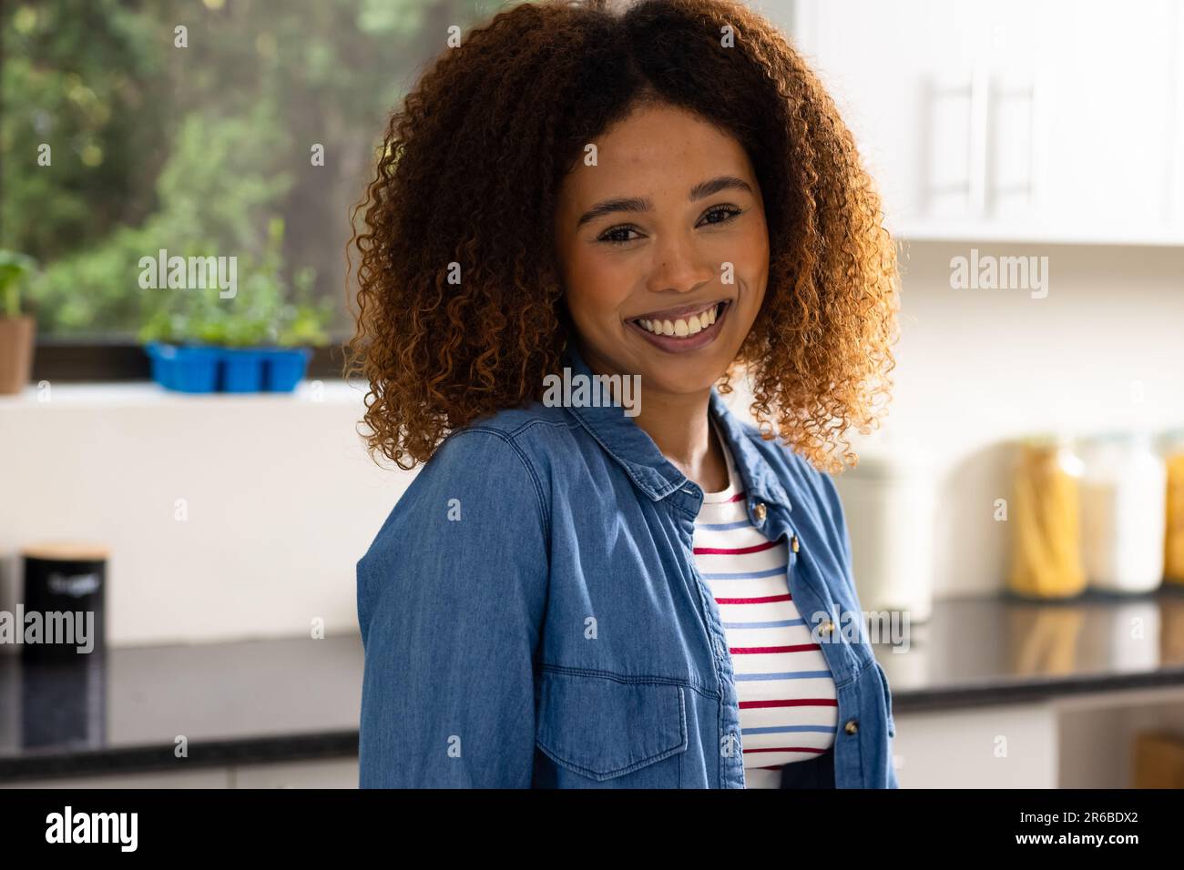 Retrato de mujer biracial feliz con el pelo rizado sonriendo en la cocina Foto de stock