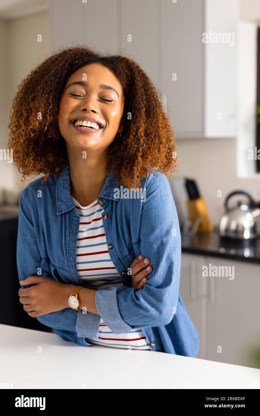 Retrato de mujer biracial feliz con el pelo rizado riendo en la cocina Foto de stock