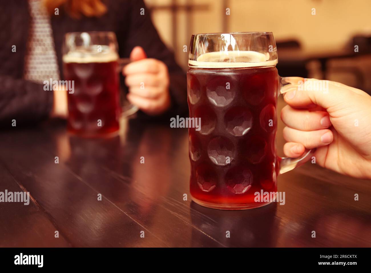 En un acogedor pub, dos amigos están disfrutando de una refrescante taza de cerveza ligera. Sus manos levantan los vasos de cerveza stein y suavemente los tintinean Foto de stock